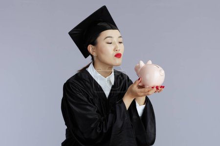 Foto de Feliz joven graduado estudiante asiático mujer con alcancía contra gris fondo. - Imagen libre de derechos