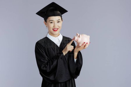 Photo for Smiling young female asian graduate student with piggy bank isolated on grey. - Royalty Free Image