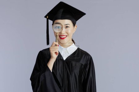 Foto de Feliz moderno graduado estudiante asiático mujer con lupa aislado en gris. - Imagen libre de derechos