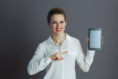 Photo for Smiling elegant 40 years old business woman in white blouse showing tablet PC blank screen isolated on gray background. - Royalty Free Image