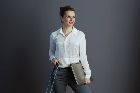 Foto de Mujer de negocios moderna y pensativa de mediana edad en blusa blanca con computadora portátil y bolsa de carro contra fondo gris. - Imagen libre de derechos