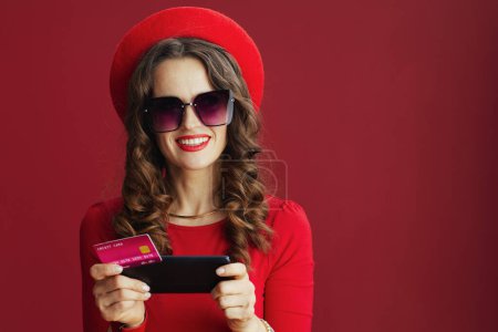 Photo for Happy Valentine. happy stylish woman with long wavy hair in red dress and beret isolated on red background with credit card and smartphone buying on internet. - Royalty Free Image