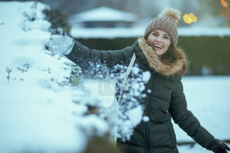 Photo for Smiling modern woman in green coat and brown hat outdoors in the city park in winter with mittens and beanie hat playing with snow. - Royalty Free Image