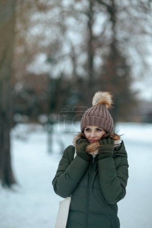 Foto de Mujer moderna y pensativa de mediana edad en abrigo verde y sombrero marrón al aire libre en el parque de la ciudad en invierno con manoplas y gorro sombrero. - Imagen libre de derechos