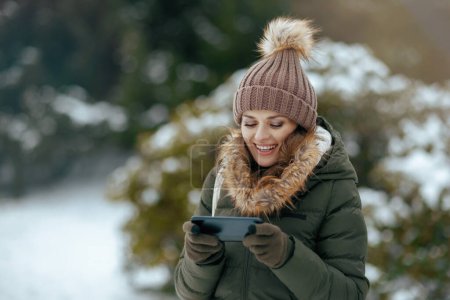 Photo for Smiling modern 40 years old woman in green coat and brown hat outdoors in the city park in winter with mittens and beanie hat using smartphone applications. - Royalty Free Image