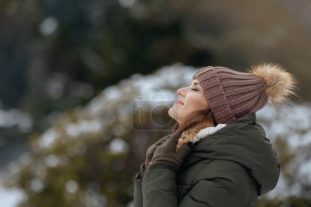 Téléchargez les photos : Heureuse femme moderne de 40 ans en manteau vert et chapeau brun à l'extérieur dans le parc de la ville en hiver avec mitaines et bonnet. - en image libre de droit