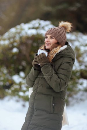 Téléchargez les photos : Femme d'âge moyen moderne souriante en manteau vert et chapeau brun à l'extérieur dans le parc de la ville en hiver avec mitaines, tasse de latte de soja et chapeau de bonnet. - en image libre de droit