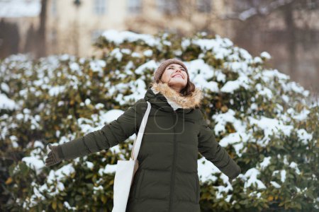 Foto de Sonriente mujer moderna en abrigo verde y sombrero marrón al aire libre en el parque de la ciudad en invierno con manoplas y gorro sombrero cerca de ramas nevadas. - Imagen libre de derechos