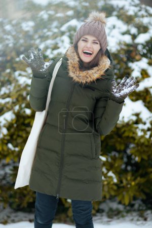 Foto de Feliz mujer moderna de mediana edad en abrigo verde y sombrero marrón al aire libre en el parque de la ciudad en invierno con manoplas y gorro sombrero cerca de ramas nevadas. - Imagen libre de derechos