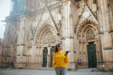 Photo for Smiling modern solo tourist woman in yellow blouse in Prague Czech Republic having walking tour, using smartphone and walking. - Royalty Free Image