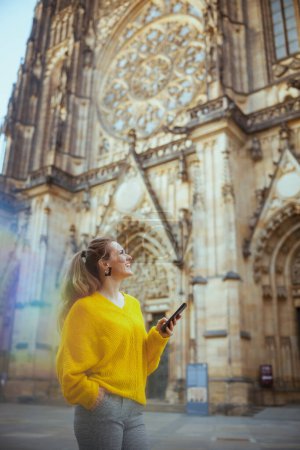 Téléchargez les photos : Voyageuse d'âge moyen souriante en chemisier jaune à Prague République tchèque explorer les attractions, en utilisant un smartphone et la marche. - en image libre de droit