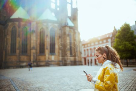 Téléchargez les photos : Heureuse voyageuse élégante en chemisier jaune et imperméable à Prague République tchèque avec smartphone ayant excursion. - en image libre de droit