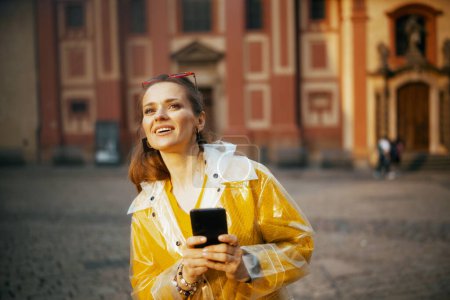 Photo for Surprised stylish middle aged traveller woman in yellow blouse and raincoat in Prague Czech Republic using smartphone. - Royalty Free Image