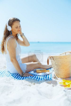 Foto de Mujer elegante y relajada en la costa del océano con bolsa de paja, auriculares y toalla a rayas. - Imagen libre de derechos