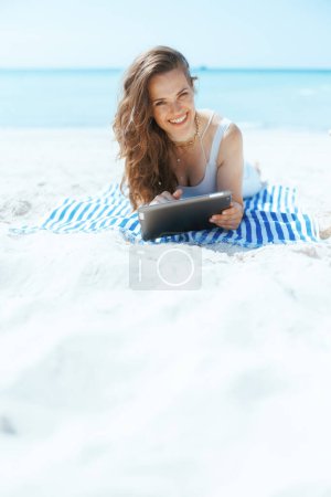 Photo for Smiling stylish woman on the seacoast with striped towel using app on tablet PC. - Royalty Free Image