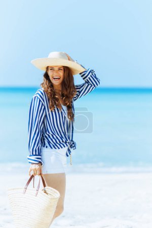 Téléchargez les photos : Sourire élégante femme âgée de 40 ans sur le bord de la mer avec sac de paille et chapeau de paille. - en image libre de droit