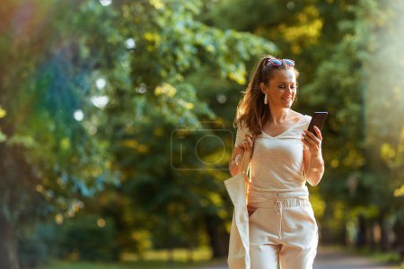 Foto de Hora de verano. mujer de moda sonriente en camisa blanca con bolsa de mano usando aplicaciones de teléfonos inteligentes y caminando en el parque de la ciudad. - Imagen libre de derechos