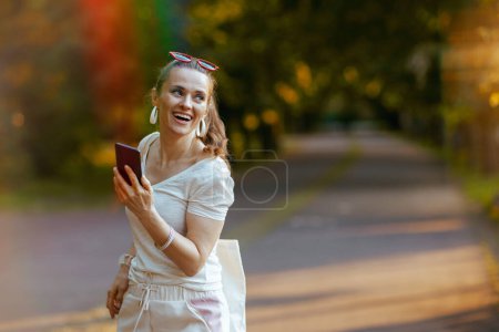 Foto de Hora de verano. mujer elegante sonriente en camisa blanca con teléfono inteligente y bolso de mano caminando en el parque de la ciudad. - Imagen libre de derechos