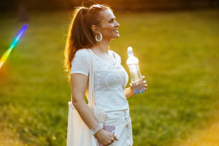 Téléchargez les photos : L'heure d'été. heureuse femme élégante en chemise blanche avec sac fourre-tout eau potable dans la prairie à l'extérieur dans la nature. - en image libre de droit