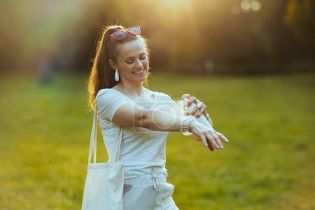 Foto de Hora de verano. sonriente mujer con estilo en camisa blanca con bolsa de mano usando spray repelente en el prado al aire libre. - Imagen libre de derechos