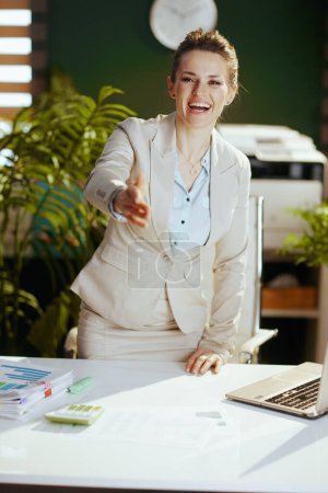 Foto de Retrato de mujer contable moderna feliz de 40 años en un traje de negocios ligero en la oficina verde moderna que da la mano para un apretón de manos. - Imagen libre de derechos