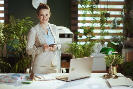 Foto de Retrato de la mujer propietaria moderna feliz de la pequeña empresa en un traje de negocios ligero en la oficina verde moderna con el ordenador portátil usando aplicaciones de teléfonos inteligentes. - Imagen libre de derechos