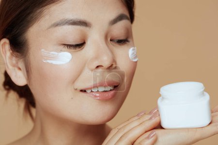Photo for Young asian female with facial cream jar and facial cream on face on beige background. - Royalty Free Image