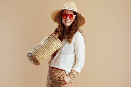 Téléchargez les photos : Vacances à la plage. Femme souriante moderne de 40 ans en chemisier blanc et short isolé sur beige avec sac en paille, chapeau en paille et lunettes de soleil. - en image libre de droit
