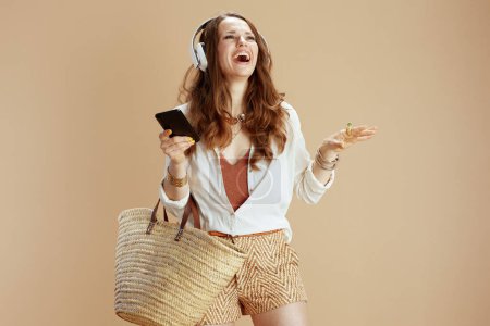 Photo for Beach vacation. smiling stylish middle aged woman in white blouse and shorts isolated on beige with straw bag and headphones using smartphone. - Royalty Free Image