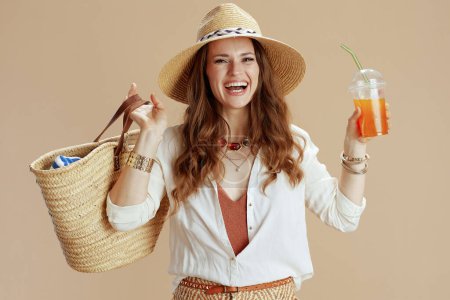 Photo for Beach vacation. happy elegant 40 years old housewife in white blouse and shorts on beige background with straw bag, carrot smoothie and straw hat. - Royalty Free Image