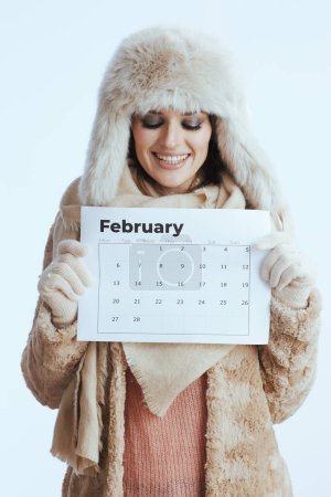 Photo for Smiling modern 40 year old woman in winter coat and fur hat isolated on white in white gloves with february calendar. - Royalty Free Image