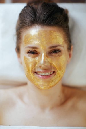 Photo for Healthcare time. Upper view of smiling modern 40 years old woman in massage cabinet with golden cosmetic mask on face laying on massage table. - Royalty Free Image