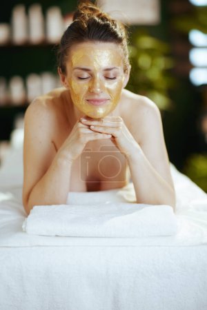 Foto de Tiempo de atención médica. mujer moderna relajada en el salón de spa con máscara cosmética dorada en la cara que pone en la mesa de masaje. - Imagen libre de derechos