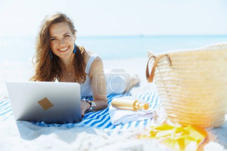 Photo for Happy modern middle aged woman on the seashore with straw bag, laptop and striped towel. - Royalty Free Image