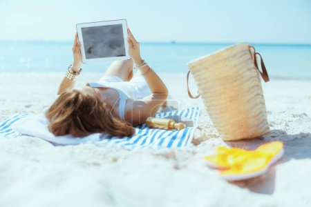 Téléchargez les photos : Détendue élégante femme âgée de 40 ans sur le littoral avec sac de paille et serviette rayée écran blanc tablette numérique. - en image libre de droit