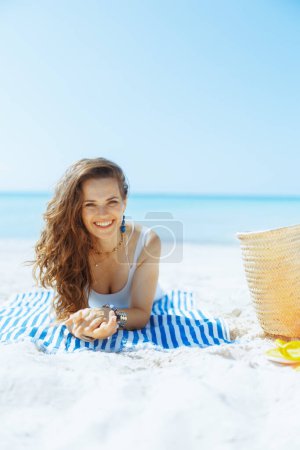 Téléchargez les photos : Femme élégante souriante sur le bord de la mer avec sac de paille et serviette rayée. - en image libre de droit