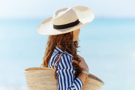 Foto de Relajado elegante hembra en la costa con bolsa de paja y sombrero de paja. - Imagen libre de derechos