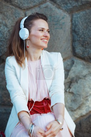 Téléchargez les photos : Femme moderne heureuse en robe rose et veste blanche dans la ville à l'écoute de la musique avec écouteurs contre mur de pierre. - en image libre de droit