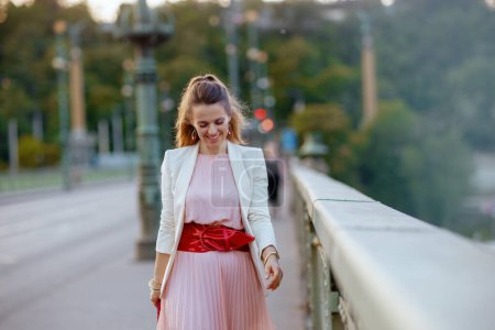 Photo for Smiling trendy middle aged woman in pink dress and white jacket in the city on the bridge. - Royalty Free Image