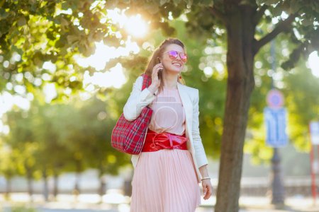Foto de Mujer elegante sonriente en vestido rosa y chaqueta blanca en la ciudad con bolsa roja y gafas de sol hablando en un teléfono inteligente. - Imagen libre de derechos