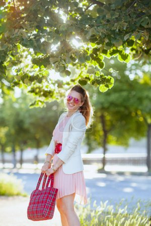 Téléchargez les photos : Femme élégante et souriante en robe rose et veste blanche dans la ville avec sac rouge et lunettes de soleil. - en image libre de droit