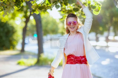 Téléchargez les photos : Heureuse élégante femme de 40 ans en robe rose et veste blanche dans la ville avec des lunettes de soleil. - en image libre de droit