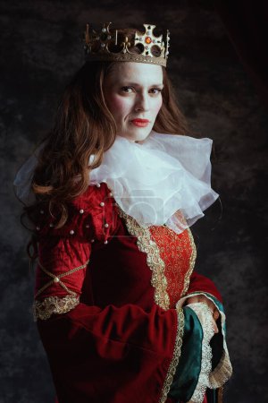 Téléchargez les photos : Reine médiévale en robe rouge avec col blanc et couronne sur fond gris foncé. - en image libre de droit