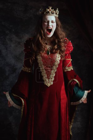 Foto de Furiosa reina medieval en vestido rojo con maquillaje blanco y corona sobre fondo gris oscuro. - Imagen libre de derechos