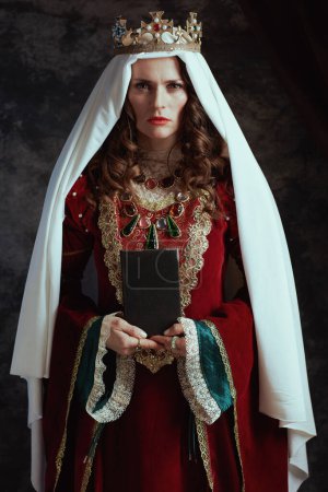 Téléchargez les photos : Reine médiévale en robe rouge avec livre, voile et couronne sur fond gris foncé. - en image libre de droit
