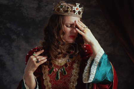 Téléchargez les photos : Reine médiévale stressée en robe rouge avec couronne ayant mal à la tête sur fond gris foncé. - en image libre de droit