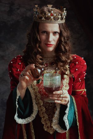 Téléchargez les photos : Reine médiévale insidieuse en robe rouge avec couronne versant du poison dans le gobelet sur fond gris foncé. - en image libre de droit