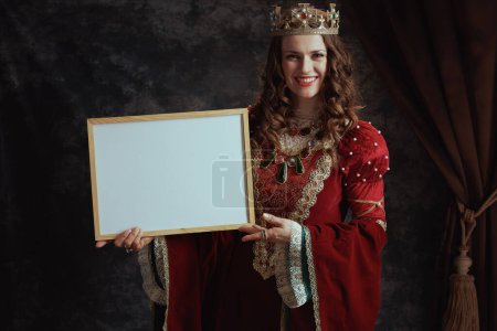 Foto de Sonriente reina medieval en vestido rojo con corona mostrando tablero en blanco sobre fondo gris oscuro. - Imagen libre de derechos