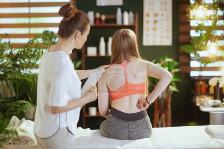 Photo for Healthcare time. Seen from behind female medical massage therapist in massage cabinet with clipboard and teenage client checking clients back condition during checkup. - Royalty Free Image