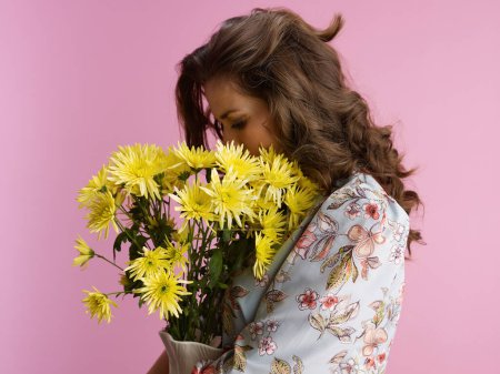 Photo for Relaxed young female with long wavy brunette hair with yellow chrysanthemums flowers in vase isolated on pink. - Royalty Free Image